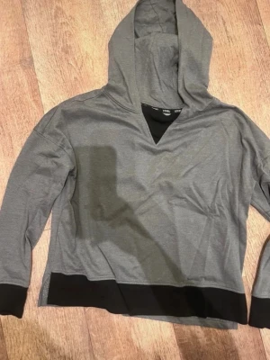 Grå hoodie med svarta detaljer - Grå hoodie med svarta muddar och svart kant nertill. Modellen har en luva och v-ringad hals med svart inslag. Perfekt för en avslappnad och stilren look.