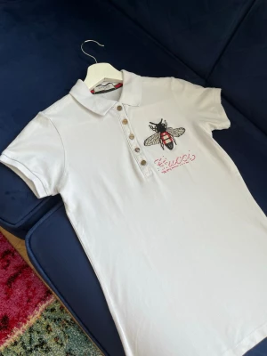 Vit pikétopp med bi och knappar - Vit pikétopp från  med korta ärmar, klassisk krage och knappslå framtill. På bröstet finns en broderad bi  i rött. Toppen är gjord i mjuk bomull och har en slim passform.