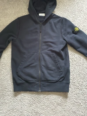 hoodie från Stone Island - Säljer en hoodie från Stone Island med dragkedja och klassisk logopatch på vänster ärm. Hoodien har stora fickor framtill och ribbade muddar. Materialet är mjukt och bekvämt, perfekt för en avslappnad look.