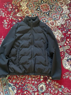 Zara Cardigan, Svart med krage - Snygg svart pufferjacka med hög krage och dragkedja framtill. Jackan har två sidofickor och elastisk nederkant för extra komfort. Perfekt för kalla dagar och ger en stilren look. Materialet är vadderat och håller värmen bra.