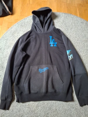 Svart LA Dodgers hoodie Champion XL - Snygg svart hoodie från Champion med LA Dodgers tryck i blått på bröstet och fickan. Stor luva och känguruficka framtill. Detalj med palmer på ena ärmen. Perfekt för dig som gillar sportig streetstyle och vill representera Dodgers.