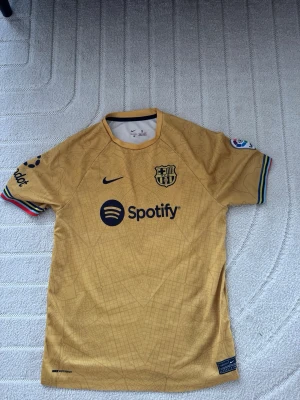 FC Barcelona bortatröja Lewandowski - Snygg guld/gul FC Barcelona bortatröja från Nike med Lewandowski och nummer 9 på ryggen. Tröjan har Spotify-logga på bröstet, La Liga-märke på ärmen och färgglada detaljer vid ärmsluten. Tillverkad i lätt och ventilerande material, perfekt för fotboll eller träning.
