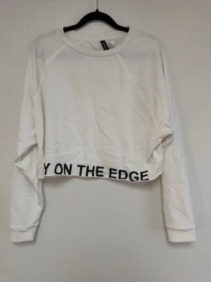 Vit croppad sweatshirt H&M - Vit croppad sweatshirt från H&M med svart text 'STAY ON THE EDGE' längs nederkanten. Tröjan har långa ärmar, rund halsringning och är gjord i mjukt bomullsmaterial. Perfekt för en avslappnad och trendig look. ( tänk på jag har hund i hemmet) 
