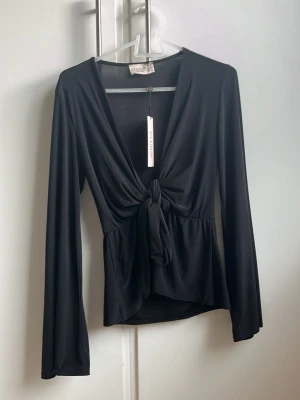 Svart långärmad topp med knyt - Trendig svart långärmad topp från Gina Tricot med djup v-ringning och snygg knytning framtill. Tillverkad i mjukt och stretchigt syntetmaterial som ger en smickrande passform. Perfekt för att lyfta vilken outfit som helst. Prislappen är på helt ny oanvänd 