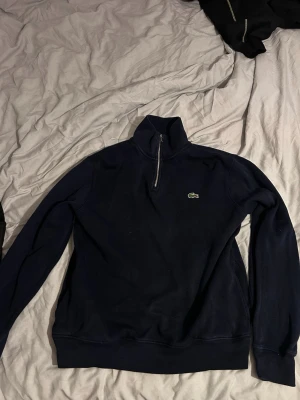 Mörkblå half zip tröja Lacoste - Mörkblå långärmad tröja från Lacoste med half zip och hög krage. Klassisk krokodillogga på bröstet. Tillverkad i mjukt material som passar perfekt till lager på lager. Ribbstickade muddar vid ärmslut och nederkant.