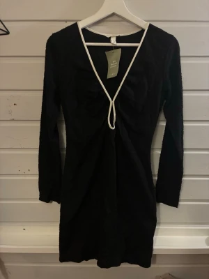 Svart långärmad klänning H&M - Svart långärmad klänning från H&M med vit kant längs den djupa v-ringningen. Tillverkad av 59% återvunnen polyester. Kort modell med figurnära passform och enkel, stilren design som passar perfekt till olika tillfällen.