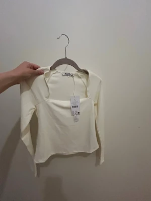 Krämvit långärmad topp från NA-KD -  Krämvit långärmad topp från NA-KD med fyrkantig urringning och figurnära passform. 