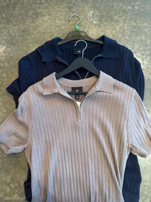 Ribbad polotopp med dragkedja H&M - Två ribbade polotoppar från H&M, en i marinblått och en i beige. Båda har korta ärmar, krage och en snygg dragkedja framtill. Perfekt till jeans eller kjol för en trendig look.