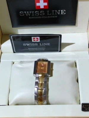 Swiss Line guld och silver klocka - Stilren klocka från Swiss Line med rektangulär urtavla i guldton och armband i både guld och silver. Urtavlan har minimalistisk design med små guldfärgade markeringar och Swiss Line-logga. Klockan har safirglas och quartz-urverk. Behöver nytt batteri 