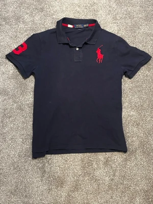 Marinblå pikétröja från Ralph Lauren - Klassisk marinblå pikétröja från Ralph Lauren med röd broderad logga på bröstet och siffran 3 på ärmen. Tröjan har krage, knappar framtill och röda detaljer på insidan av kragen. Perfekt för en sportig och stilren look. Den här pucken är lite mindre i storleken vilket gör den perfekt passande för personer som är mellan 170 cm och 190 cm vid minsta fundering var vänligen att meddela frågor. Med vänliga hälsningar c
