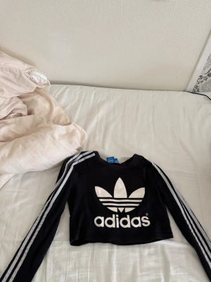 Svart croppad långärmad topp Adidas - Säljer en svart croppad långärmad topp från Adidas med vita ränder längs ärmarna och stort Adidas-tryck framtill. Perfekt för en sportig och trendig look. Rund halsringning och slim passform.