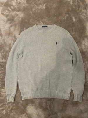 Ljusgrå stickad tröja från Polo Ralph Lauren - Snygg ljusgrå stickad tröja från Polo Ralph Lauren med rund halsringning och diskret blå logga broderad på bröstet. Tröjan har långa ärmar och ribbade muddar vid ärmslut och nederkant. Perfekt för lager på lager och enkel att matcha.