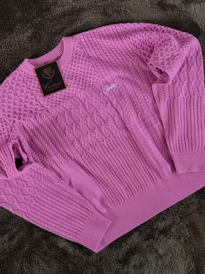 Corteiz Pink Knitted Sweater  - Säljer en rosa stickad tröja från Corteiz med flera olika stickade mönster, inklusive flätor och bikake-struktur. Tröjan har rund halsringning, ribbade muddar och broderad logga på bröstet. Perfekt för dig som gillar färg och detaljer.