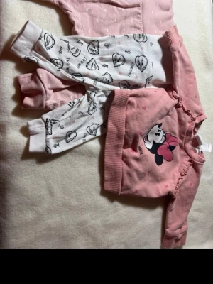 Sött babyset med Minnie Mouse - Två par mjuka byxor och en långärmad tröja för baby. Ena byxan är ljusrosa med små vita hjärtan, den andra vit med svart Minnie Mouse-tryck. Tröjan är rosa med volangdetaljer och stor Minnie Mouse-tryck framtill. Alla plagg i bomullsmix och bekväm passform.