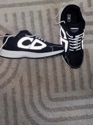Dior b30 - Säljer ett par svarta Dior sneakers med vita reflexdetaljer och stora CD-loggor på sidan. Skorna har vita snören, meshpaneler och en chunky vit sula. Perfekta för dig som vill sticka ut med en lyxig och modern look.