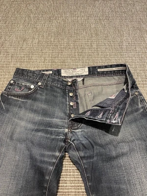 Jacob Cohen jeans  - Säljer ett par mörkblå jeans från Cohen med raka ben och snygga slitningar. Jeansen har klassiska kontrastsömmar, knäppning med knappar och detaljerade fickor med logga. Perfekta för en avslappnad och trendig look.