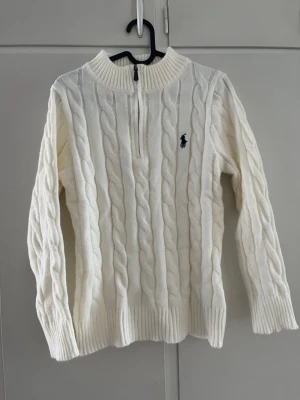 Vit kabelstickad tröja Polo Ralph Lauren - Kabelstickad vit tröja från Polo Ralph Lauren med halv dragkedja och ribbad krage. Tröjan har lång ärm, ribbade muddar och den klassiska broderade loggan på bröstet. Perfekt för dig som gillar en clean och tidlös stil. Tröjan är aldrig använd, skulle  rekommendera till dig som har XS i vanliga fall