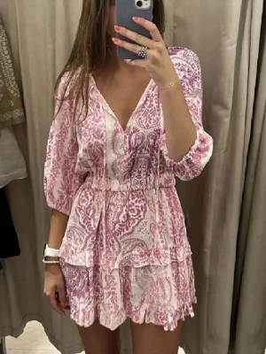 Rosa och vit kortklänning med volang - Söt kortklänning i rosa och vitt med blommigt mönster och volangdetaljer. Klänningen har v-ringning, resår i midjan och trekvartsärm. Perfekt för sommarens alla tillfällen.