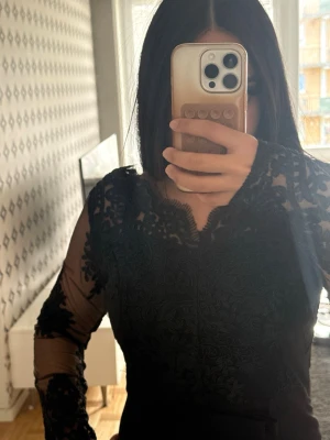 Svart lång klänning med lång ärm - Snygg svart klänning med transparenta spetsärmar och detaljerat spetsmönster  över hela framsidan. Blusen har en v-ringad halsringning med vågig kant och är tillverkad i ett tunt, elegant material som ger en lyxig känsla.