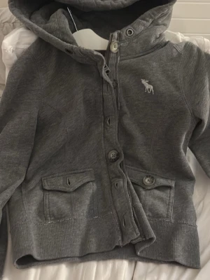 Grå hoodie från Abercrombie & Fitch - Mysig grå hoodie med knappar och dragkedja från Abercrombie & Fitch. Har två fickor framtill, ribbade muddar och en broderad älglogga på bröstet. Perfekt för chill dagar och enkel att matcha med jeans eller mjukisbyxor.