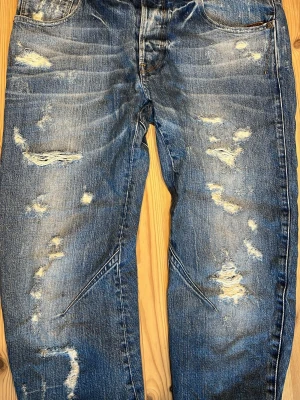 Blå slitna jeans med hål - Säljer ett par blå jeans med kraftigt slitna detaljer och flera hål framtill. Jeansen har en avslappnad passform, klassiska fem fickor och kontrastsömmar. Perfekta för en trendig och avslappnad streetstyle-look.