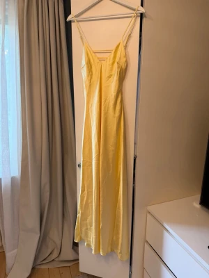 Gul långklänning från Zara - Snygg ljusgul långklänning med smala axelband och v-ringning både fram och bak. Klänningen har en elegant, slät siluett och är tillverkad i ett glansigt material som ger en lyxig känsla. Perfekt för sommarens alla tillfällen.