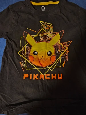 Svart Pikachu t-shirt Pokémon + shorts  - Cool svart t-shirt med stort Pikachu-tryck framtill och grafiska detaljer i gult och orange. Rund halsringning med gul insida. Perfekt för alla Pokémon-fans som vill sticka ut med en färgglad och lekfull look.