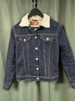 Mörkblå jeansjacka med teddykrage - Snygg mörkblå jeansjacka från Replay med vit teddykrage och kontrastsömmar. Klassisk modell med två bröstfickor med lock och knapp, samt knappstängning framtill. Perfekt för dig som vill ha en tidlös och trendig look.