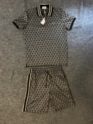Lacoste piké och shorts - Snyggt set från Lacoste med pikétröja och matchande shorts. Setet är svart och grått med ett grafiskt diamantmönster, svarta ribbade detaljer och vita ränder på shortsen. Perfekt för en sportig och trendig look.