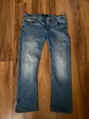 Blå raka jeans - Säljer ett par klassiska blå jeans med rak passform och fem fickor. Jeansen har en snygg tvätt med ljusa slitningar på framsidan och baksidan. 