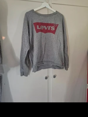Grå sweatshirt från Levi's - Klassisk grå sweatshirt från Levi's med stort rött logotryck på bröstet. Tröjan har rund halsringning, långa ärmar och ribbade muddar vid ärmslut och nederkant. Perfekt för en avslappnad och stilren look.