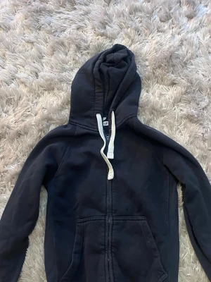 Svart zip-hoodie med vita snören - Klassisk svart hoodie från Junkyard med dragkedja framtill och stora fickor. Utrustad med huva och vita, breda dragsnören. Tillverkad i mjukt material som känns skönt mot huden. Perfekt för en avslappnad och stilren look.