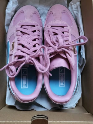 Rosa Puma sneakers med blå detalj - Snygga sneakers från Puma i en fräsch rosa färg med blå sidodetalj och klassisk gummisula. Skorna har snörning, guldfärgad Puma-logga på sidan och är tillverkade i mocka och läder. Perfekta för dig som vill sticka ut med färg på fötterna.