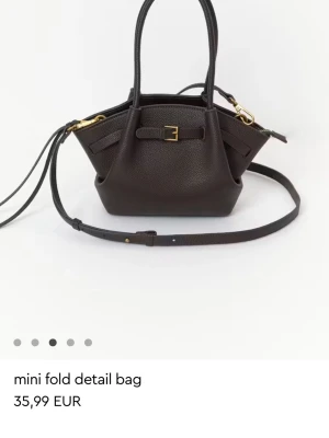 Mörkbrun mini handväska med spänne - Snygg mörkbrun mini handväska i konstläder med guldfärgade metalldetaljer och justerbar axelrem. Väskan har en unik vikt design och dekorativt spänne framtill. Perfekt storlek för det viktigaste och enkel att bära både i handen och över axeln.