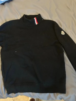 Moncler tröja  - Svart stickad tröja från Moncler med hög krage och röd, vit och blå randdetalj vid halsen. Tröjan har Moncler-logga på ena ärmen och ribbstickade muddar vid ärmslut och nederkant. Perfekt för dig som gillar stilrena och exklusiva plagg.