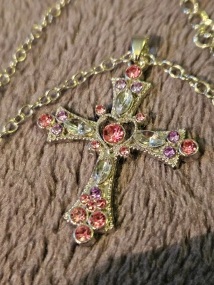 Korshalsband med rosa och lila stenar - Snyggt halsband med ett stort korsformat hänge i silverfärg, dekorerat med glittrande rosa och lila stenar samt klara pärlliknande detaljer. Hänget har ett hjärta i mitten och kedjan är i matchande silverton. Perfekt för dig som gillar statement-smycken.