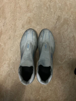 Adidas F50 vita fotbollsskor - Säljer ett par Adidas F50 fotbollsskor i vitt med blå detaljer. Skorna har en strömlinjeformad design utan snörning och är tillverkade i syntetmaterial för lätt vikt och bra bollkontroll. Perfekta för dig som vill ha snabbhet och stil på planen.