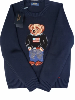 Mörkblå stickad tröja Polo Bear - Mörkblå stickad tröja från Polo Ralph Lauren med ikonisk Polo Bear på bröstet. Björnen har amerikansk flagga på tröjan, jeans och bruna skor. Tröjan har rund halsringning och ribbstickade muddar. Perfekt för dig som gillar klassisk preppy stil.