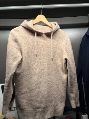 Beige hoodie i ull mix  - Säljer en beige hoodie med dragsko och långa ärmar. Tröjan är stickad i en mjuk ullmix och har ribbade muddar vid ärmslut och nederkant. Perfekt för en chill och avslappnad stil.