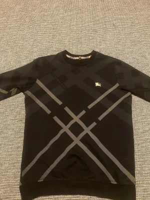 Svart Burberry tröja med rutmönster - Snygg svart tröja från Burberry med klassiskt rutmönster i grått och diskret Burberry-logga på bröstet. Tröjan har rund halsringning och långa ärmar. Perfekt för dig som gillar stilrena och exklusiva plagg.