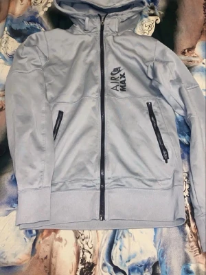 Nike tech - Ljusblå hoodie från Nike Air Max med svart logga på bröstet. Tröjan har dragkedja framtill, två svarta dragkedjefickor och en stor huva. Ribbade muddar vid ärmslut och nederkant. Materialet är mjukt och bekvämt, perfekt för chill eller träning.