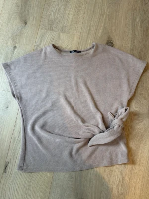 Beige topp med knyt från Zara S - Säljer en beige topp från Zara i mjukt material. Toppen har korta ärmar, rund halsringning och en snygg knytdetalj i sidan som ger en cool touch. Perfekt att styla med jeans eller kjol för en avslappnad look. Storlek S