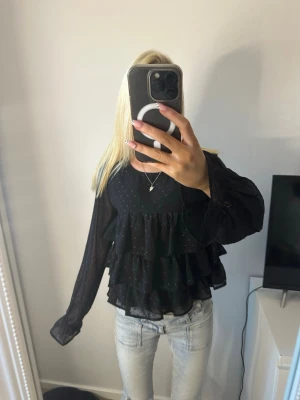 Svart volangblus med prickar - ⚠️GÅR INTE ATT KNÄPPA⚠️ Trendig svart blus med volanger i flera lager och små diskreta prickar. Blusen har transparenta långärmade ärmar och en luftig passform som ger en cool och avslappnad look. Perfekt att styla med jeans för en snygg outfit.
