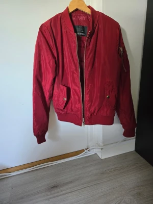 Röd bomberjacka från ONLY - Snygg röd bomberjacka från ONLY med klassisk passform. Jackan har dragkedja framtill, två fickor med tryckknappar och en extra ficka med dragkedja på ärmen. Ribbstickade muddar vid ärmslut och nederkant. Perfekt för dig som vill ha en stilren och trendig look.