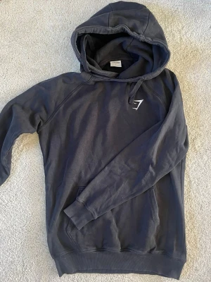 Svart hoodie från Gymshark - Snygg svart hoodie från Gymshark med vit logga på bröstet. Klassisk modell med huva och dragsnören samt en stor magficka. Tillverkad i mjukt bomullsmaterial som känns skönt mot huden. Perfekt för en avslappnad och sportig stil.