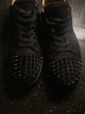 Svarta Louboutin sneakers med nitar - Säljer ett par svarta sneakers från Christian Louboutin med coola svarta nitar över tån och den klassiska röda sulan. Skorna har snörning och är tillverkade i textil och läder. Perfekta för dig som vill sticka ut med en edgy och lyxig stil.