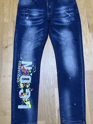  jeans - Säljer ett par blå jeans tryck i vitt, gult och rött på vänster ben. Jeansen har fem fickor, raka ben och slitna detaljer framtill. Materialet är klassisk denim med stretch för skön passform.