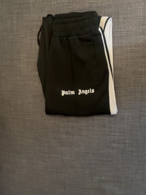 Palm Angels svarta mjukisbyxor - Svarta mjukisbyxor från Palm Angels med vita sidoränder och logga tryckt på vänster lår. Byxorna har resår i midjan och raka ben. Perfekta för en avslappnad och trendig streetwear-look.