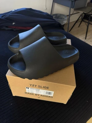Yeezy Slide tofflor från Adidas - Säljer ett par svarta YZY Slide tofflor från Adidas. De har en chunky, modern design med bred rem och räfflad sula för extra grepp. Tillverkade i mjukt och flexibelt syntetmaterial som ger en bekväm känsla. Perfekta för en avslappnad och trendig stil.