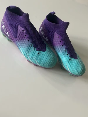 Lila och turkosa fotbollsskor TSLN - Snygga fotbollsskor från TSLN i en cool färgkombination av lila och turkos med gradient-effekt. Skorna har hög krage för extra stöd runt vristen och mönstrad ovandel för bättre bollkontroll. Perfekta för dig som vill sticka ut på planen.
